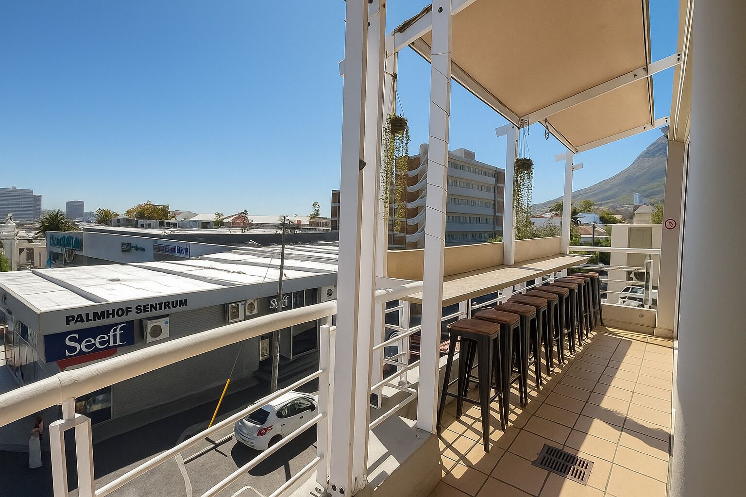 Un balcón con vista a un edificio identificado como 'PALMHOF SENTRUM' y 'Seeff', que incluye una zona de estar con taburetes y una mesa.