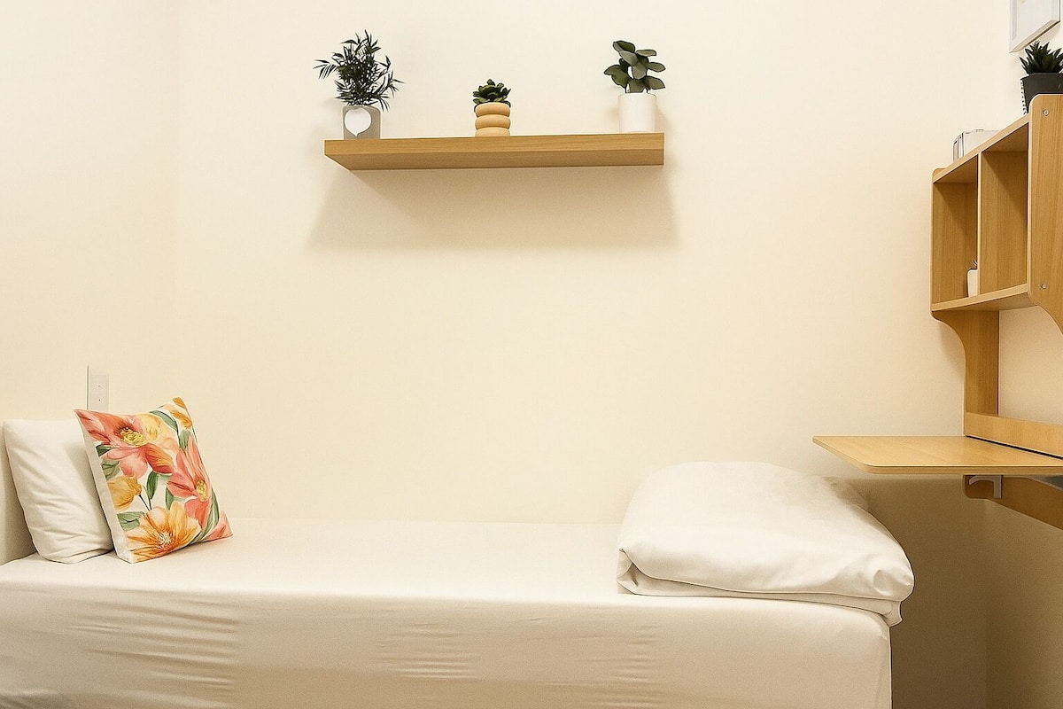 Une petite chambre avec un lit simple, au-dessus duquel se trouve une étagère en bois. Sur l’étagère, on peut voir trois plantes en pot. Sur le lit, il y a un oreiller blanc et un autre oreiller à motifs floraux.