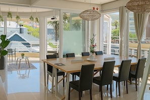 A modern dining area with a large table, chairs, hanging plants, and large windows offering a view of the outside.