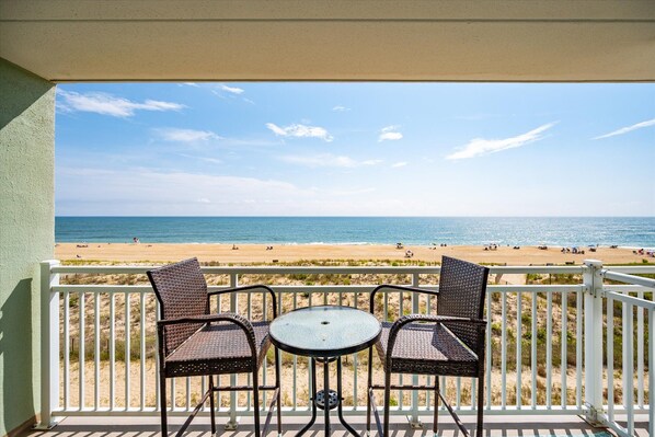 A balcony with two wicker chairs and a small round table, overlooking a beach with people and umbrellas.