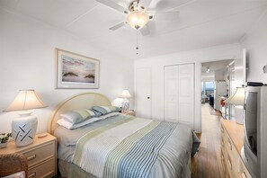 A bedroom with a bed, bedside lamps, a ceiling fan, a framed picture, and a view into an adjacent room.