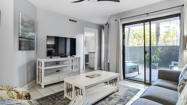 A modern living room with a flat-screen TV, white furniture, a ceiling fan, and a sliding glass door to a balcony. - First Floor Two Bedroom Two Bath Condo - 1 Block to Beach (Hilton Head Island)