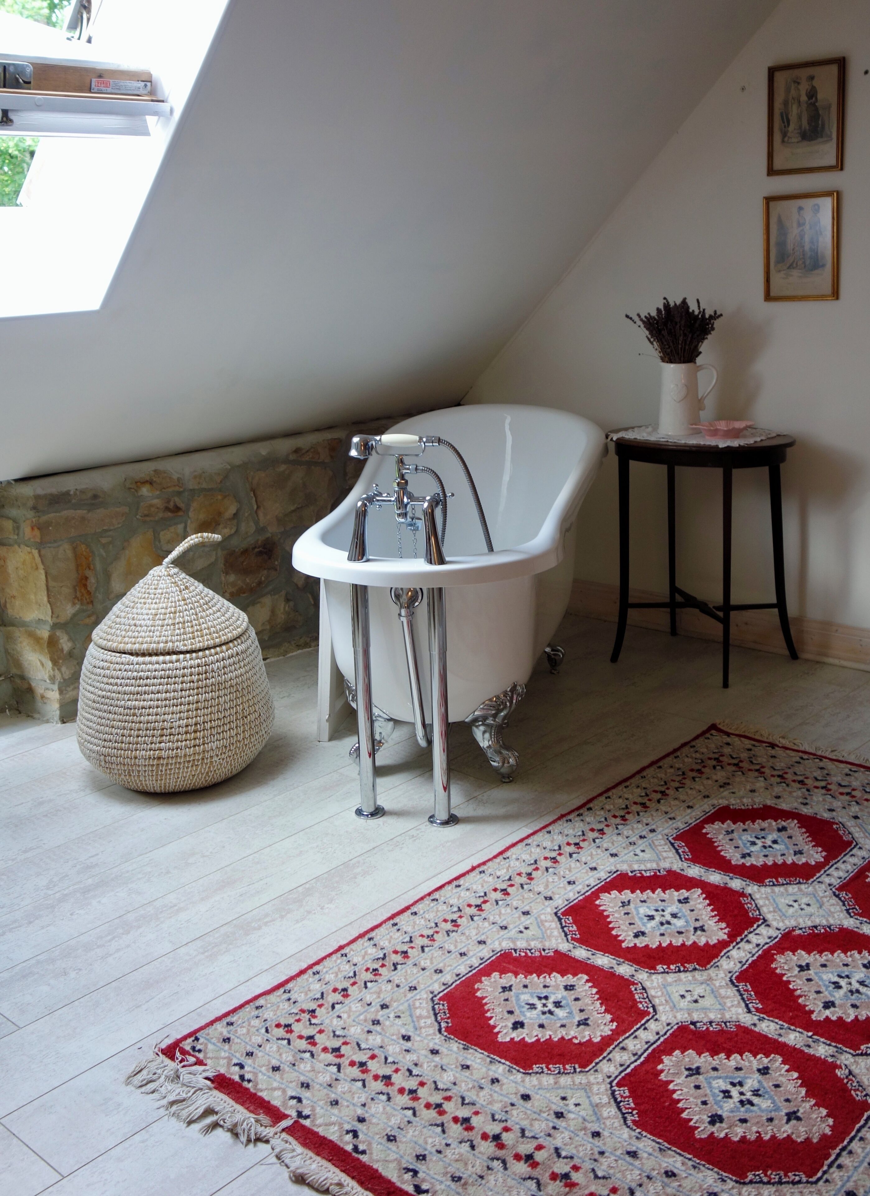 Une salle de bain avec une baignoire sur pieds, un pouf en fibres tressées, un tapis à motifs et une petite table sur laquelle est posé un vase.