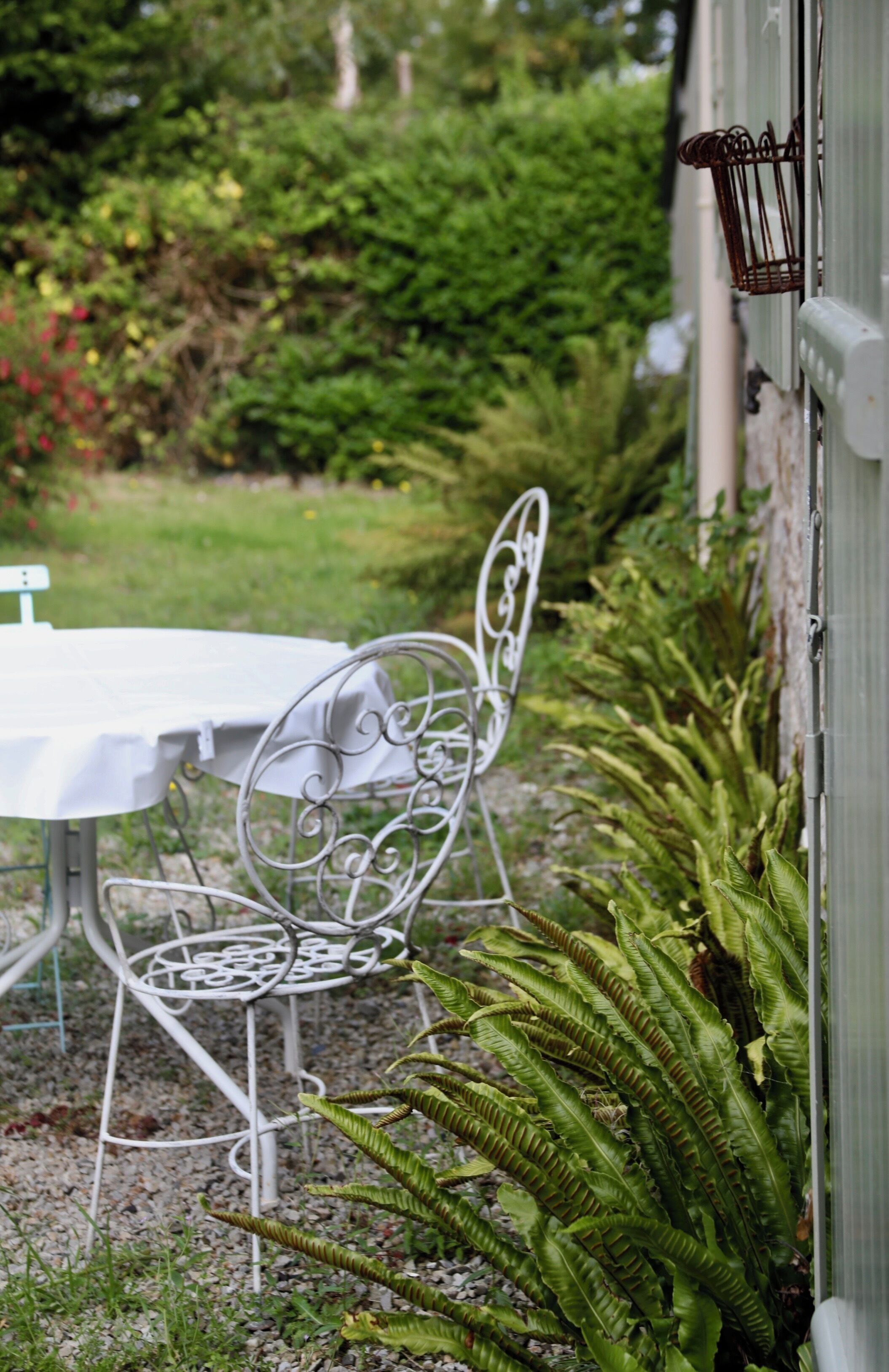 Conjunto de mesa e cadeiras de metal branco ao ar livre, rodeado por vegetação e um edifício branco.