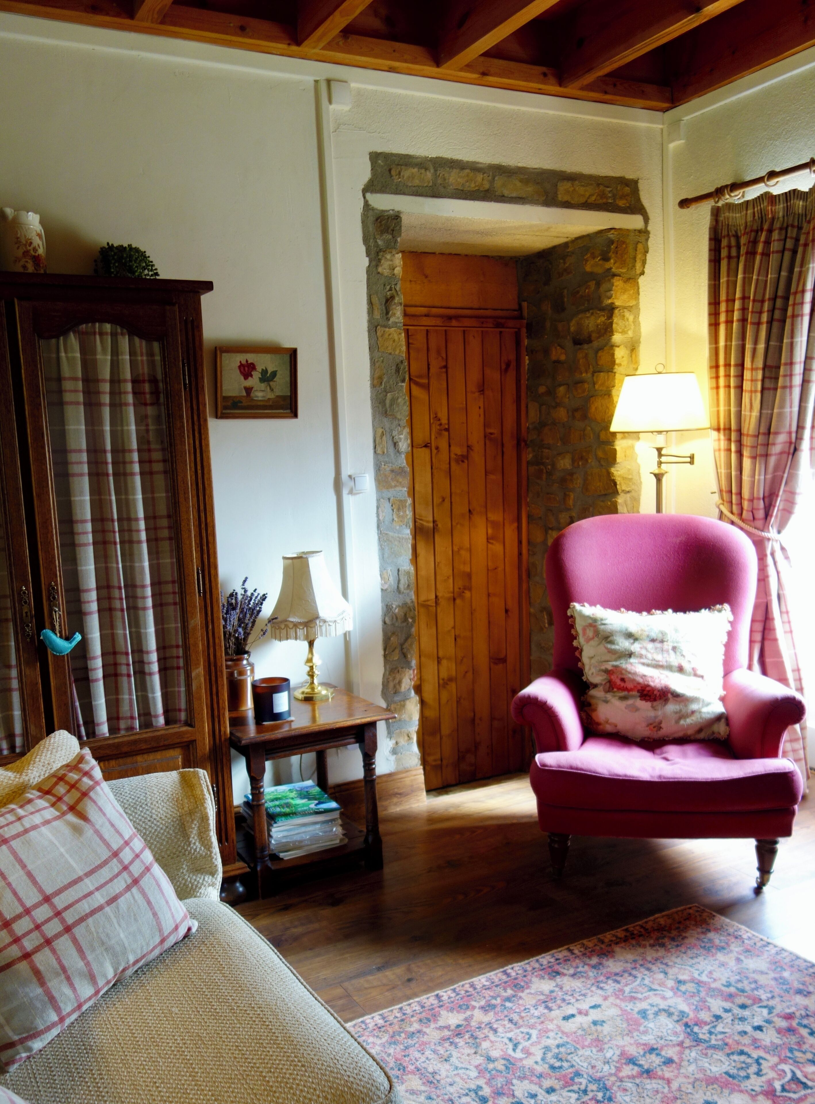 Ein gemütliches Wohnzimmer mit Holzboden, einem rosafarbenen Sessel mit Blumenkissen, einer Holzkommode und einem fensterförmigen Vorhang in Karo.