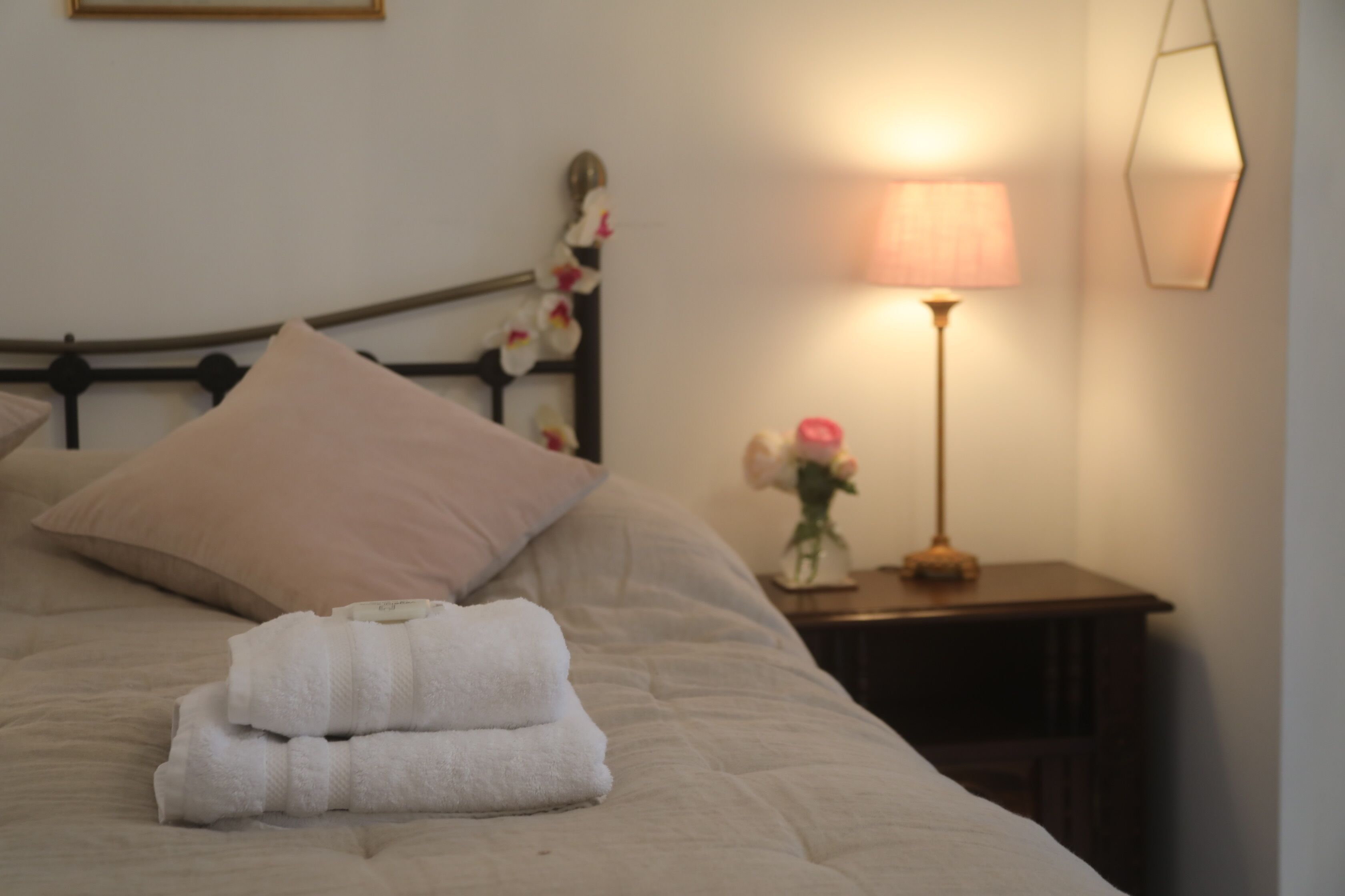 Ein ordentlich bezogenes Bett mit einer beigen Tagesdecke und Kissen, ein Nachttisch mit Lampe und einer Blumenvase, sowie eine Wandleuchte.