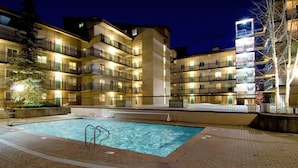 A nighttime view of a hotel with a swimming pool, multiple balconies, and a brick-paved area. - Lionshead Village 2 Bedroom With Pool, Walk to Slopes! (Vail)