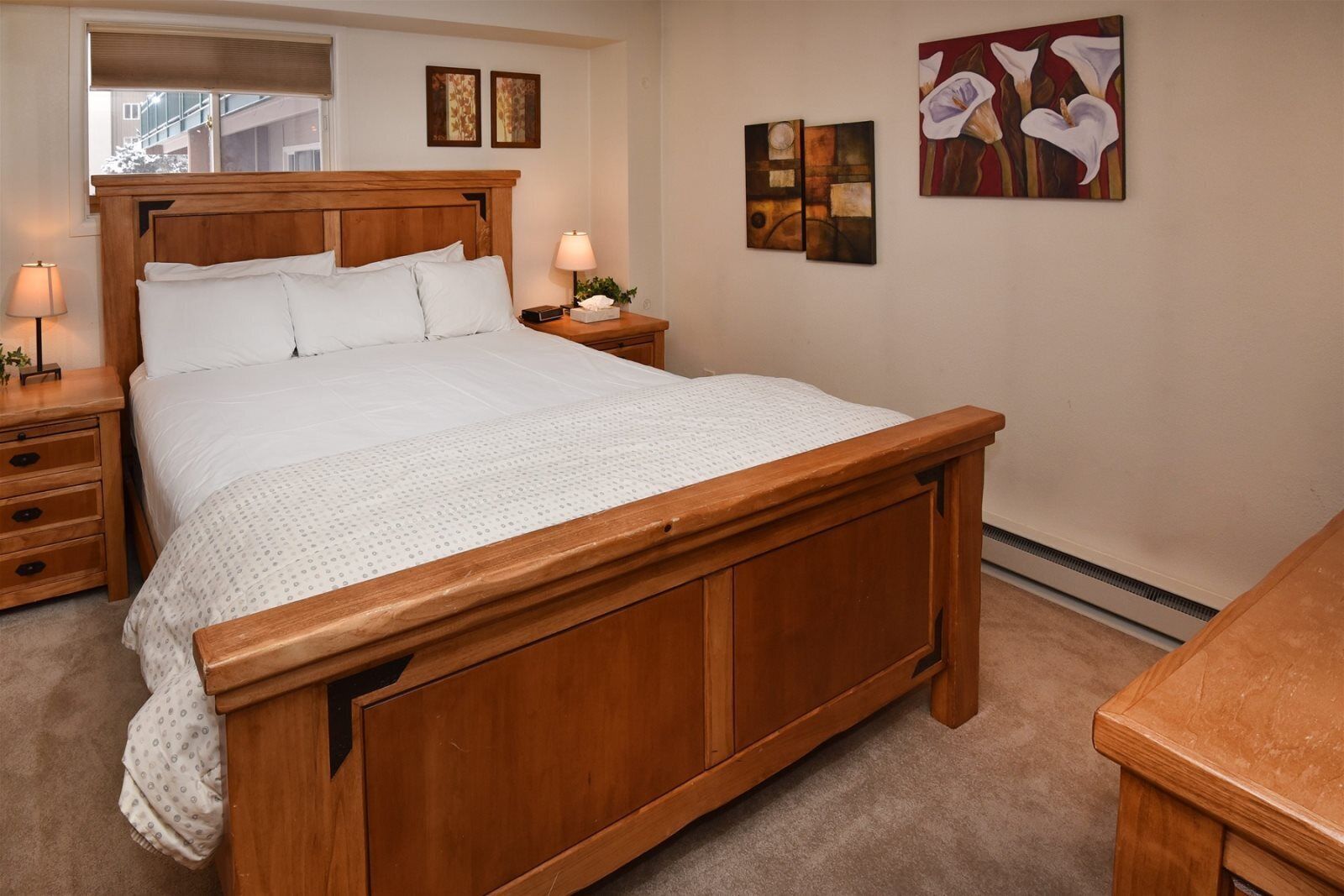 A bedroom with a wooden bed, a nightstand with a lamp, and artwork on the wall.