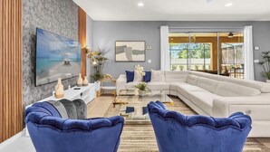 Un salon moderne avec un grand canapé d’angle, une table basse et deux fauteuils bleus. Un téléviseur à écran plat est fixé au mur, ainsi qu’un tableau et une plante en pot.