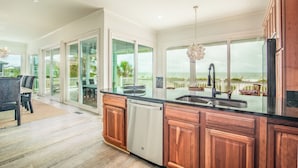A modern kitchen with wooden cabinets, a black countertop, and a view of the ocean through large windows. - The Great Escape: Oceanfront 6BR Home (Georgetown)