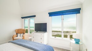 A bedroom with a bed, a dresser, a television, a chair, a lamp, and large windows with a view of the beach. - The Great Escape: Oceanfront 6BR Home (Georgetown)