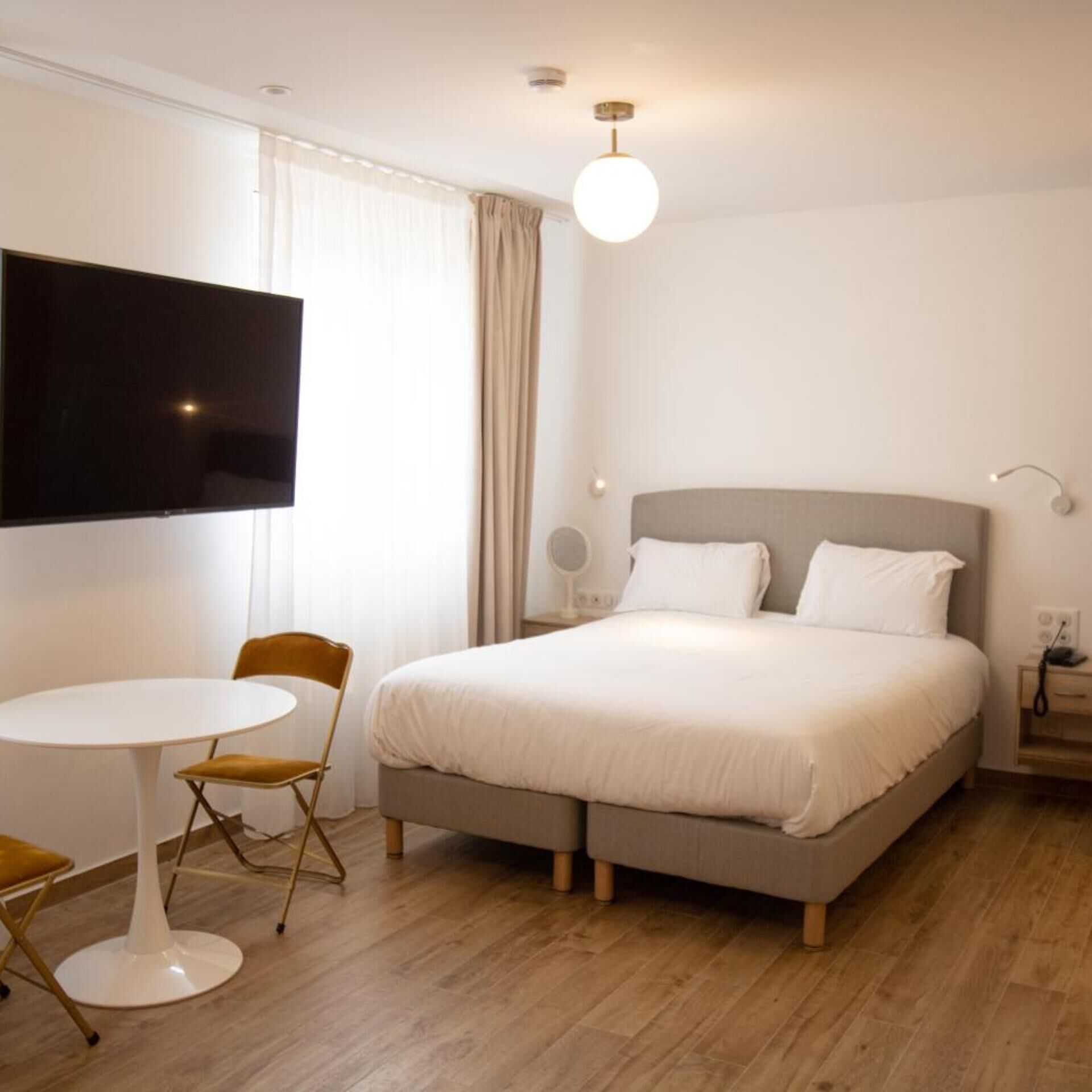 A hotel room with a bed, a television, a round table with a chair, and a window with curtains.
