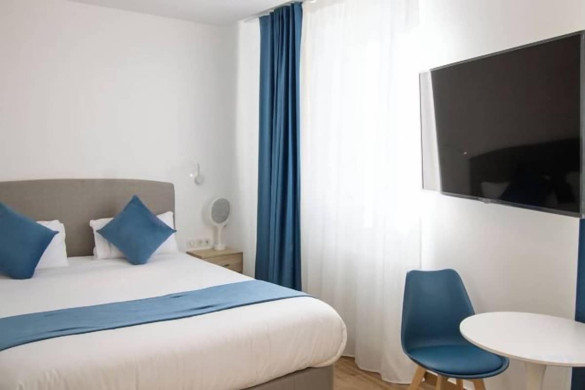 A hotel room with a bed, a bedside table, a chair, a small table, and a large window.