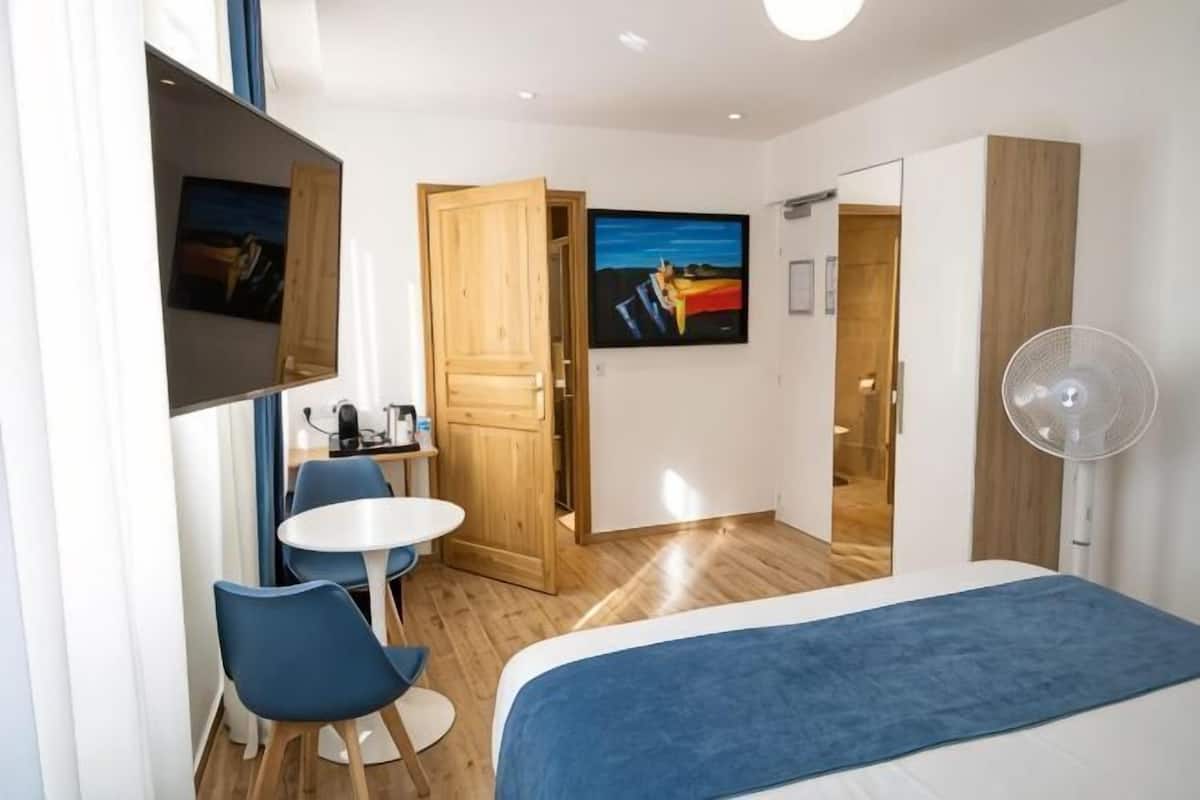 A hotel room with a bed, a desk with a chair, a TV, a fan, and a painting on the wall.