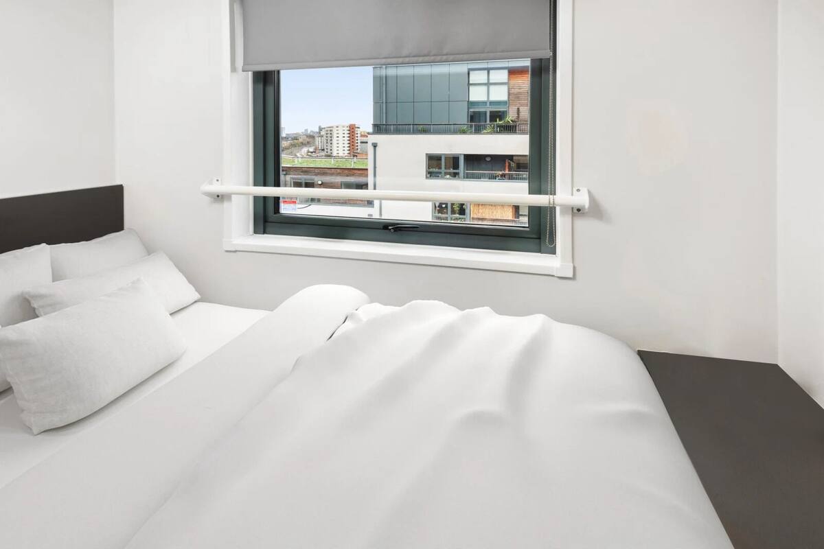 Et moderne hotelværelse med en seng, hvidt sengetøj, et vindue med udsigt til bygninger og en grå sengegavl.