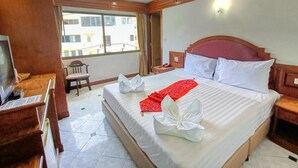 A hotel room with a large bed, wooden headboard, a desk with a chair, and a window with a view of buildings. - Phuket suite hotel  (Patong)