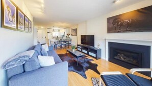 Een moderne woonkamer met een bank, een flatscreen televisie, een open haard en een eethoek op de achtergrond.