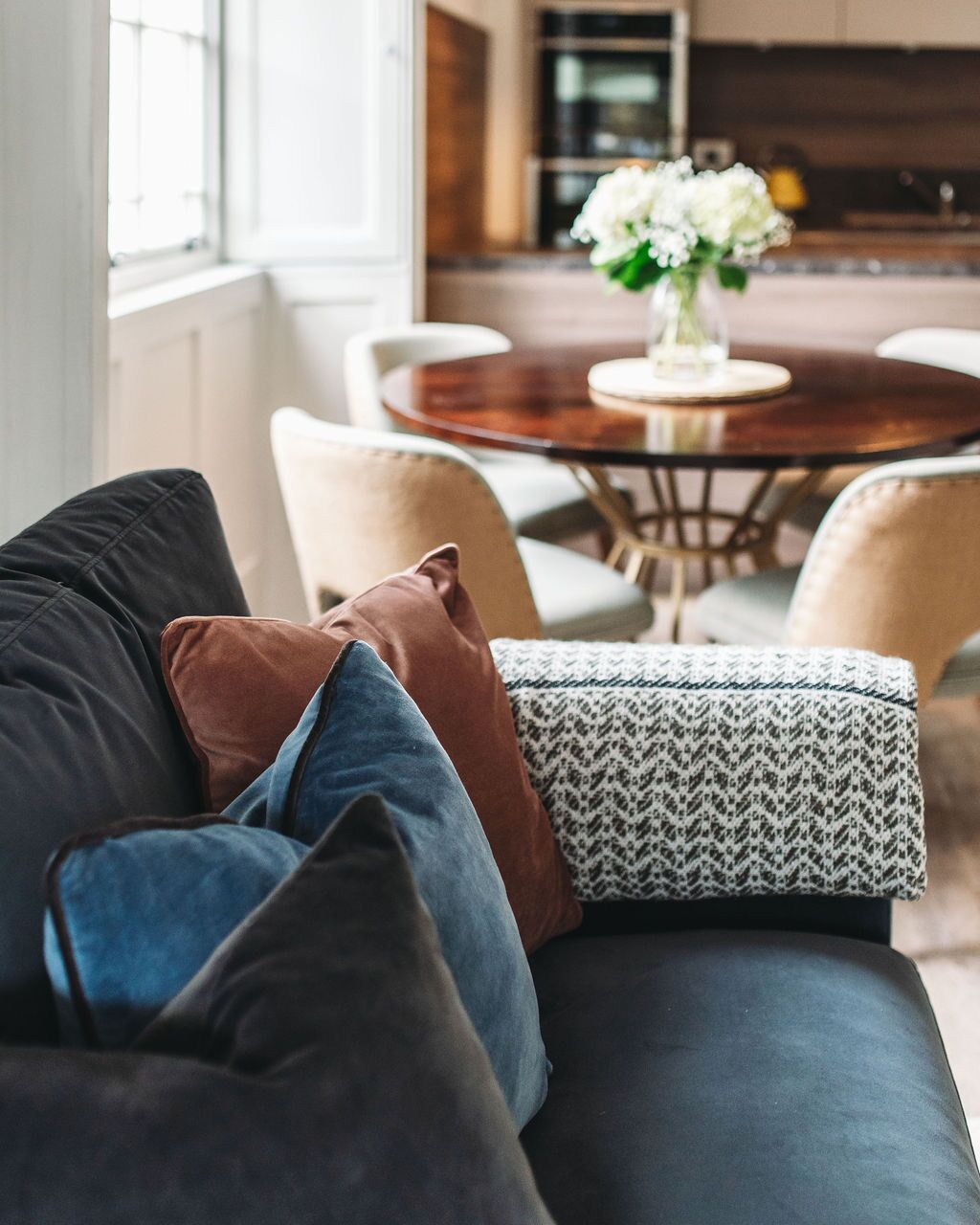 Una sala de estar moderna con un sofá gris oscuro, cojines estampados y una mesa de comedor redonda de madera con sillas.