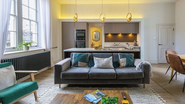 A modern living room with a grey sofa, a green armchair, and a wooden coffee table. There is a kitchen area with a stove, oven, and sink in the background. - Gorgeous, stylish, huge Georgian home 4 bed 4 bath (Clitheroe)