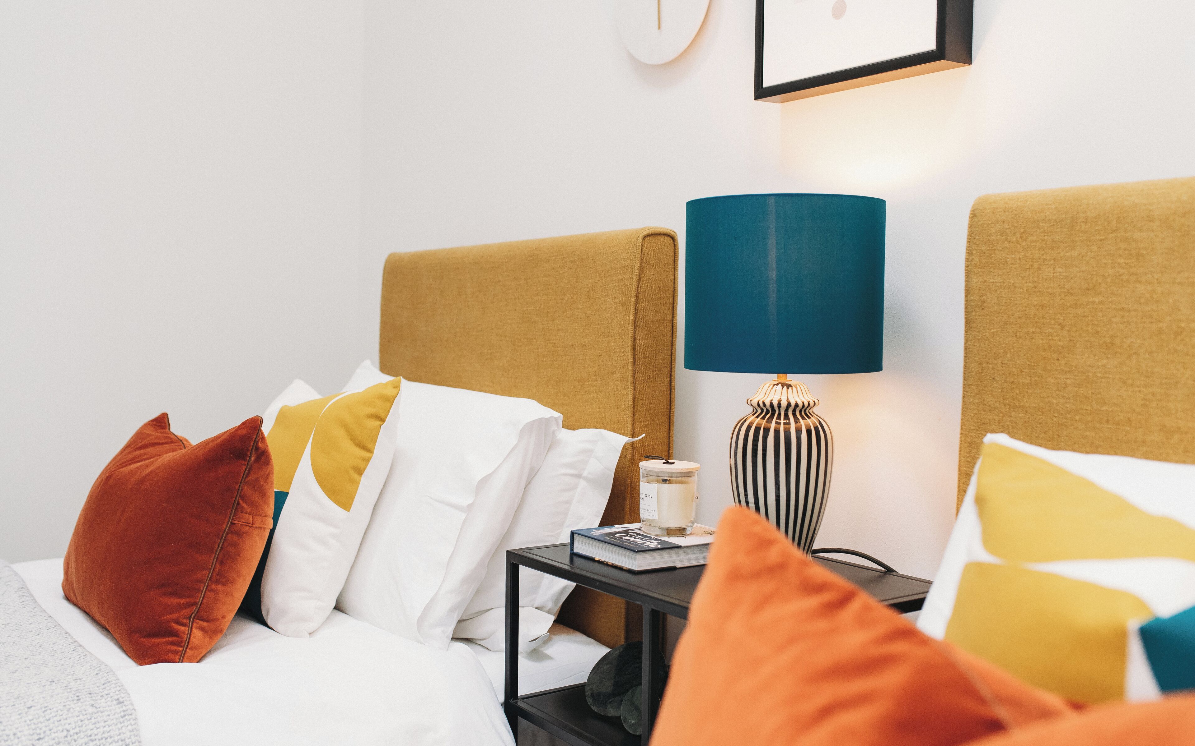 A neatly made bed with white and orange pillows, a bedside table with a lamp, and a book.