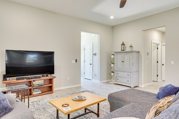 Una sala de estar con televisor de pantalla plana, una mesa de centro y un sofá. Se observa un mueble con objetos decorativos y una puerta que da a otra habitación.