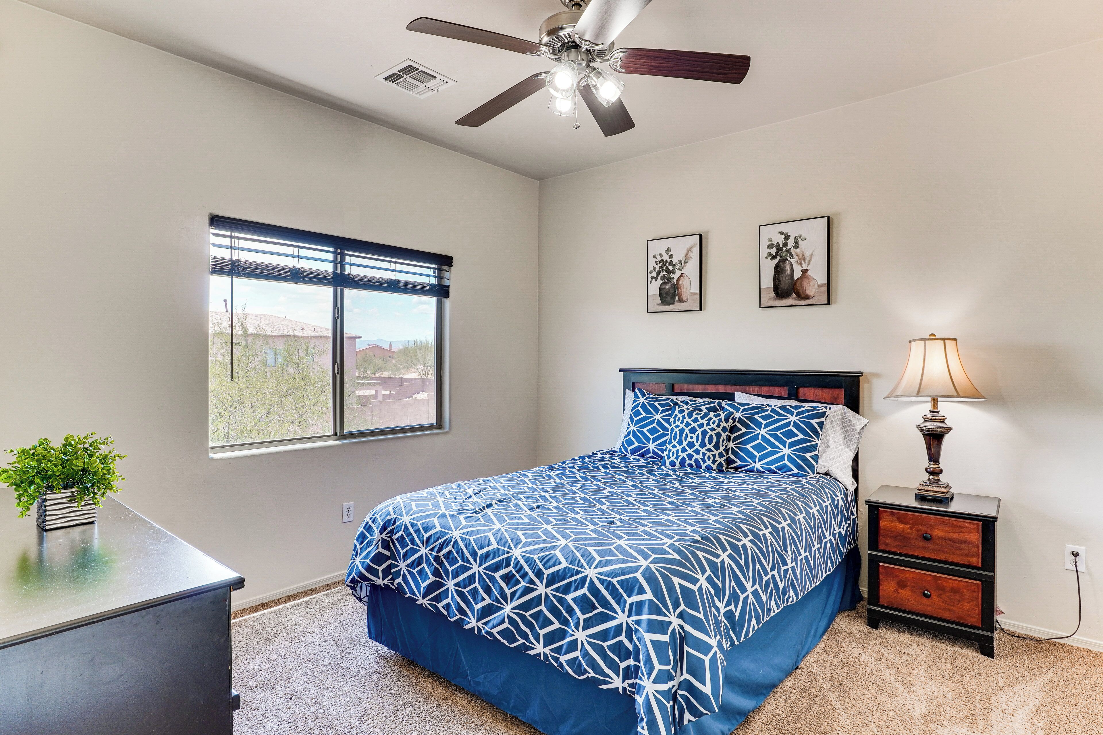 A bedroom with a bed, a nightstand, a desk, a ceiling fan, and a window with blinds.