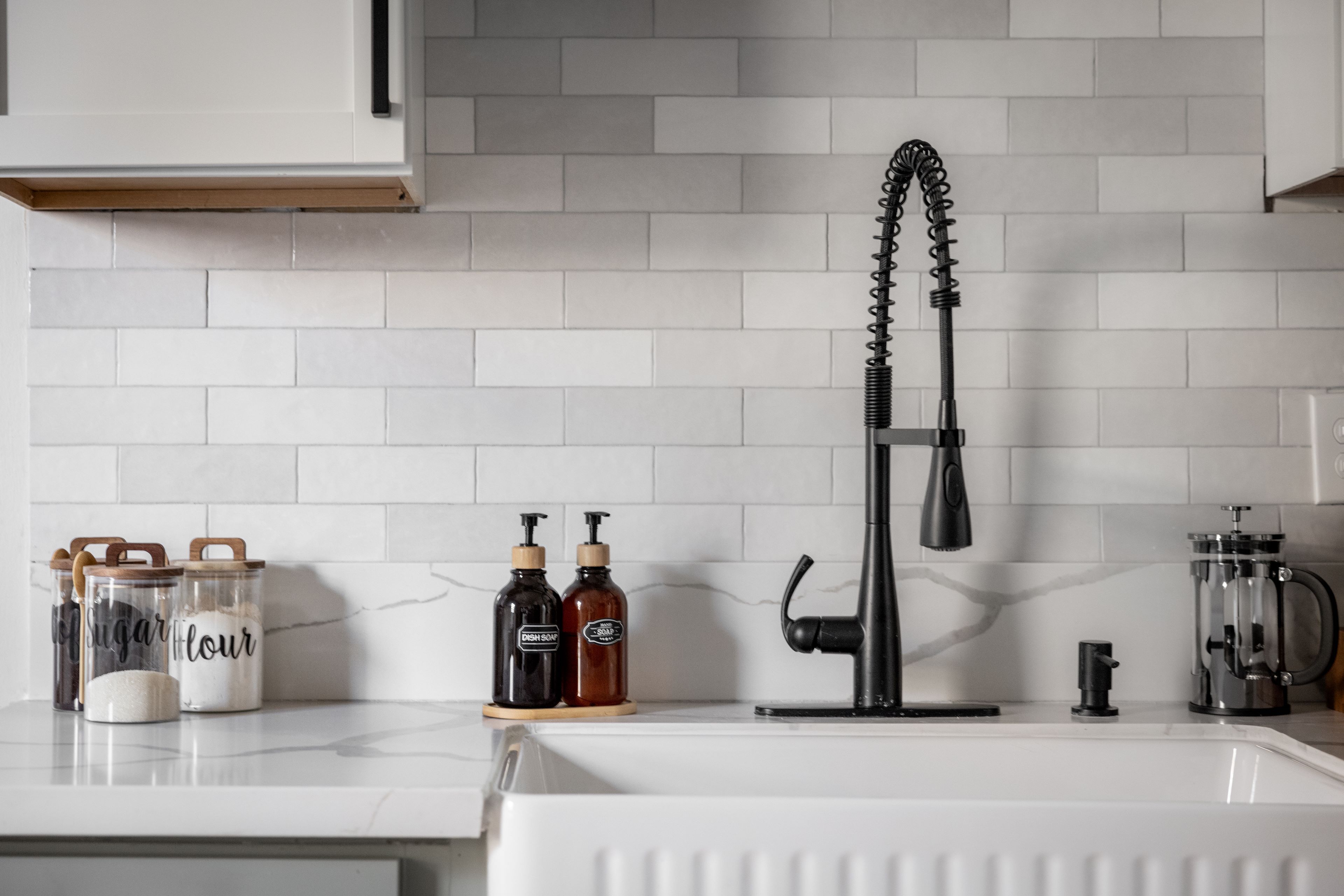 A kitchen with a white countertop, a black faucet, and jars labeled 'sugar' and 'flour'.