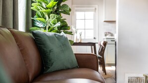 En brun lädersoffa med en grön kudde i ett rum med en stor växt, ett matbord och en vit dörr.