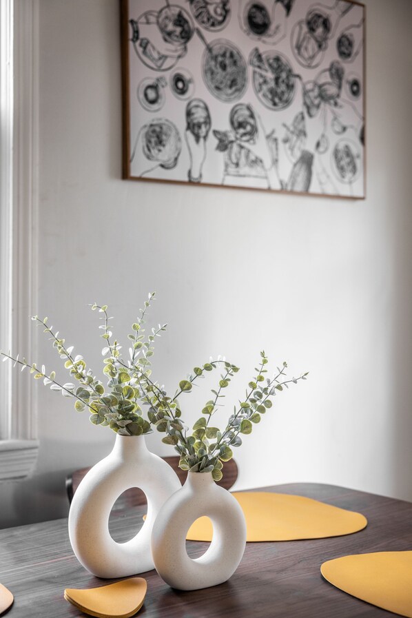 Two white vases with plants on a wooden table, a black and white abstract painting on the wall, and a yellow placemat on the table.