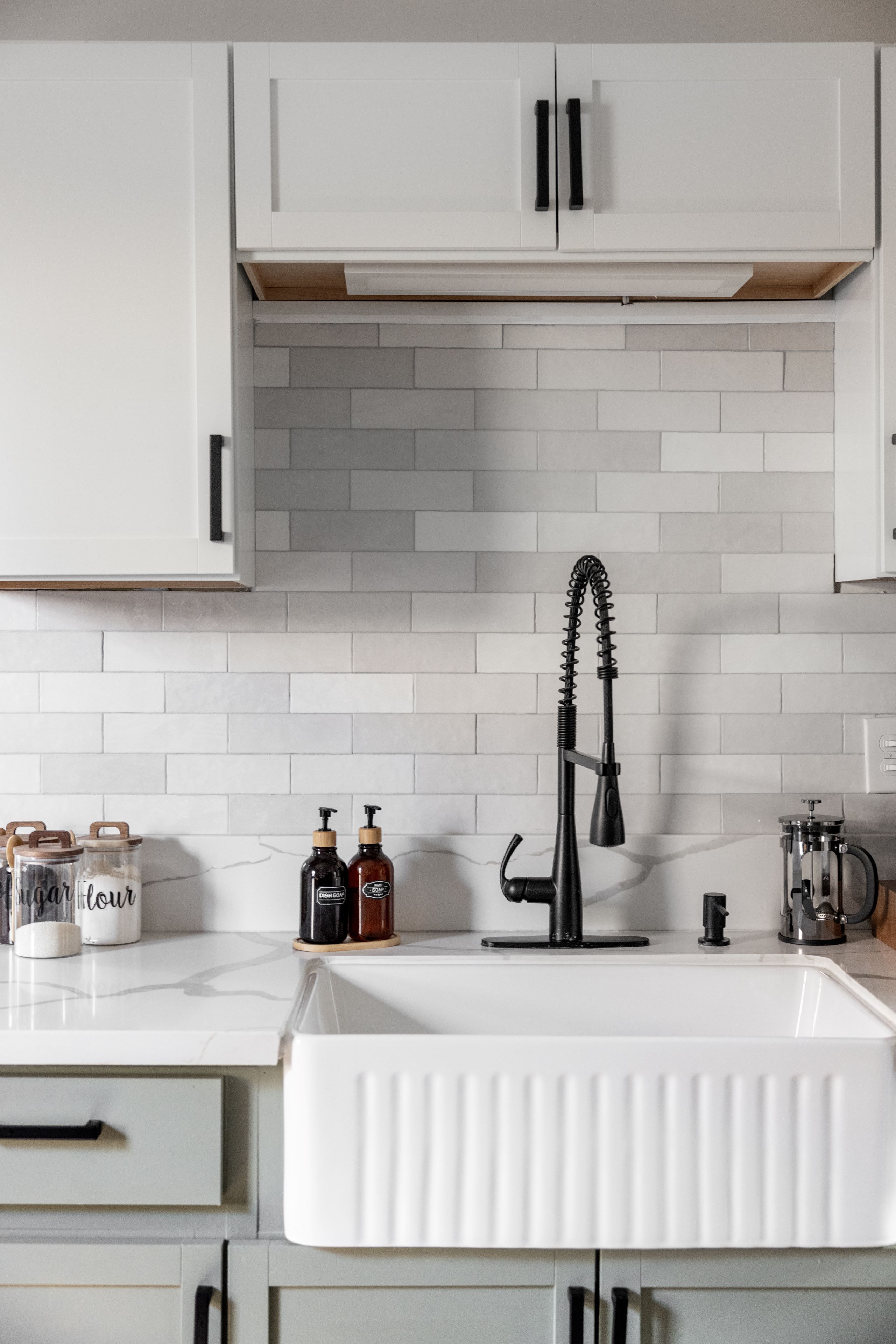 A kitchen with white cabinets, a white sink, and a backsplash. There are jars and bottles on the counter.
