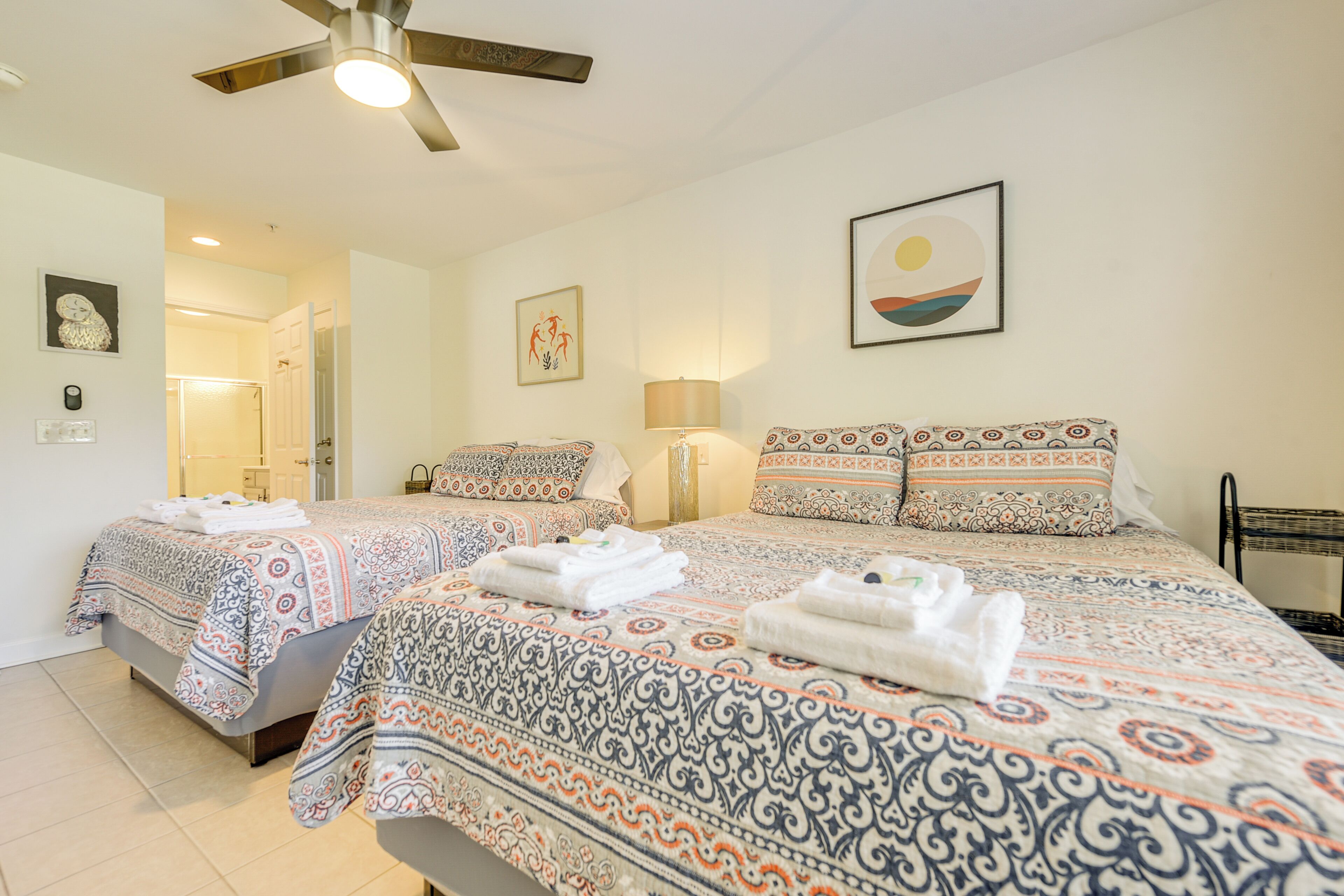Quarto de hotel com duas camas, roupa de cama estampada, ventoinha no teto e obras de arte nas paredes.