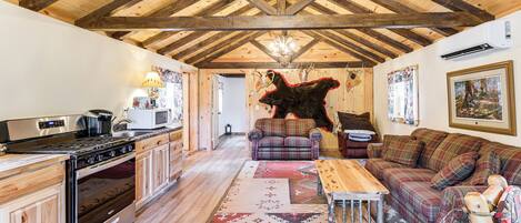 A rustic interior with a wooden ceiling, a kitchen with a stove and microwave, a plaid sofa, and a rug.