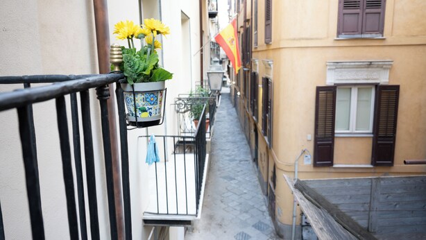 A narrow street with buildings on either side, a balcony with a hanging flower pot, and a flag on one of the buildings. - Apartment in the very center of Palermo, within walking distance of the famous 4 canti. (Palermo)