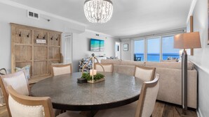 A dining area with a round table, chairs, a chandelier, and a view of the ocean through large windows. - Update 2Br Beachfront Condo - Balcony View, Pool,  and Tram. Steps to Beach! (Miramar Beach)