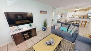 A living room with a TV, sofa, coffee table, and a kitchen in the background. - Venus 116 - Beach side 1 BR steps to the Gulf (Fort Walton Beach)