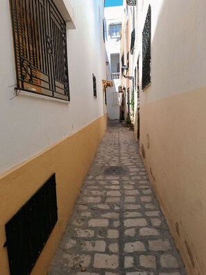 A narrow cobblestone alley with white and yellow walls, metal bars on windows, and a manhole cover. - Dar Aziz (Sousse)