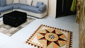 Ein Wohnzimmer mit einer Eckcouch, einem schwarzen Hocker, einem gemusterten Teppich und einer Wand mit arabischer Kalligraphie.