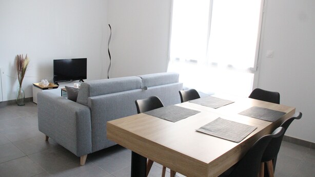 Un salon moderne avec un canapé gris, une table à manger en bois avec des chaises, un téléviseur à écran plat et un vase contenant des plantes séchées.