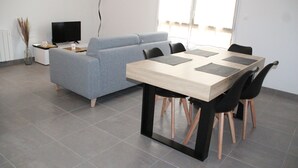 Un salon moderne avec un canapé gris, une table à manger en bois avec des chaises noires, un téléviseur à écran plat et un vase contenant des plantes séchées.