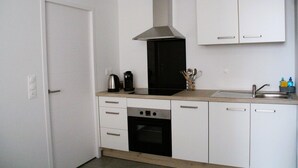 Une cuisine compacte équipée d’une cuisinière, d’un four et d’une hotte intégrée. Elle comprend des meubles blancs et un petit plan de travail avec un évier.