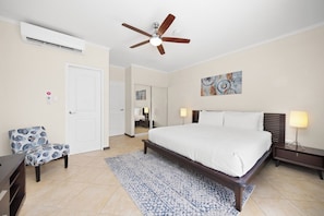 Una camera d'albergo con un letto, una ventola a soffitto, un comodino, una lampada, una sedia e un quadro appeso al muro.