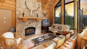 L’intérieur d’une cabane en rondins, avec une cheminée en pierre, des meubles en bois et une grande fenêtre donnant sur le paysage extérieur.