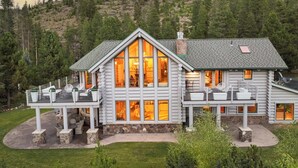 Une cabane en rondins dotée d’une grande façade vitrée, de fondations en pierre et d’une terrasse aménagée avec du mobilier d’extérieur.