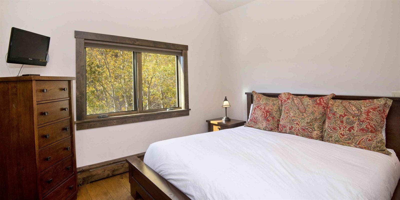 A bedroom with a bed, a dresser, a nightstand, a lamp, and a TV mounted on the wall.