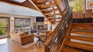 A spacious living area with a fireplace, wooden staircase, and large windows offering a view of the outdoors. - Potato Patch Luxury 4 Bedroom Townhome - Vail Point (Vail)
