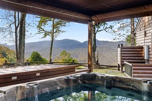 A hot tub with a mountain view, wooden deck, and a log cabin structure.