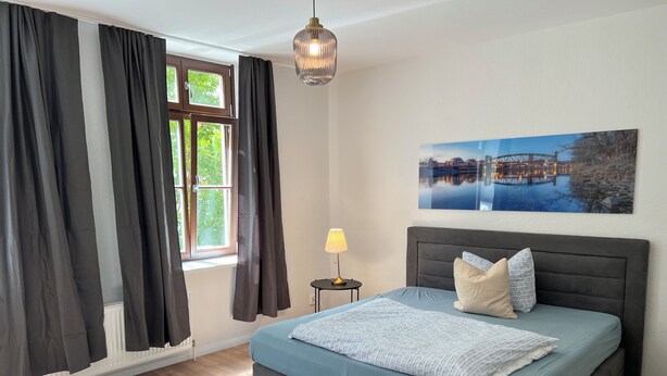 Une chambre à coucher avec un lit, une table de chevet, une lampe, une fenêtre avec des rideaux et un tableau au mur.