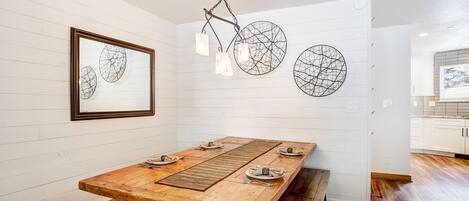 A dining area with a wooden table set for four, white paneled walls, and a framed artwork above the table.