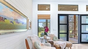 A cozy interior with two plush armchairs, a small round table, and a large painting of cows on the wall. - Wild Candy Farm - Breezeway Haus (Wimberley)