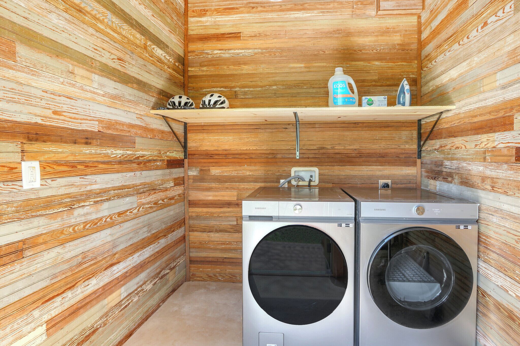 Un cuarto de lavandería con dos lavadoras Samsung, un estante con productos de limpieza y una pared de madera.